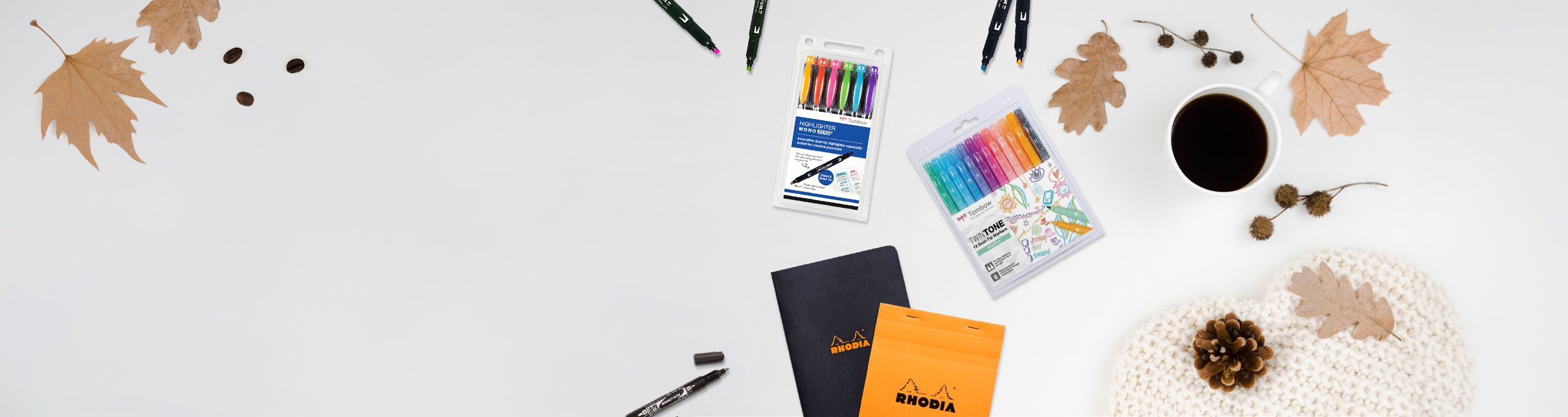 Flat lay of fall desk supplies: leaves, coffee, markers, notebooks, pouches, and a pine cone on a knitted cream blanket on a pale background.