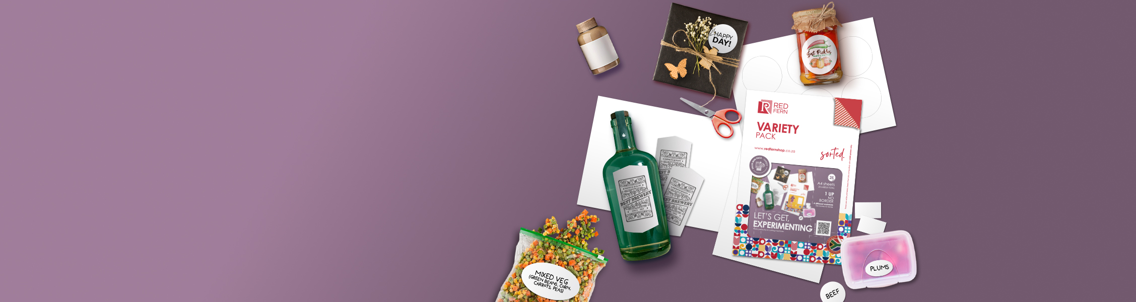 Flat lay of gift packaging items on a purple background, including a green bottle, scissors, a 'Variety Pack' brochure, jars, a bag of mixed vegetables, and labels