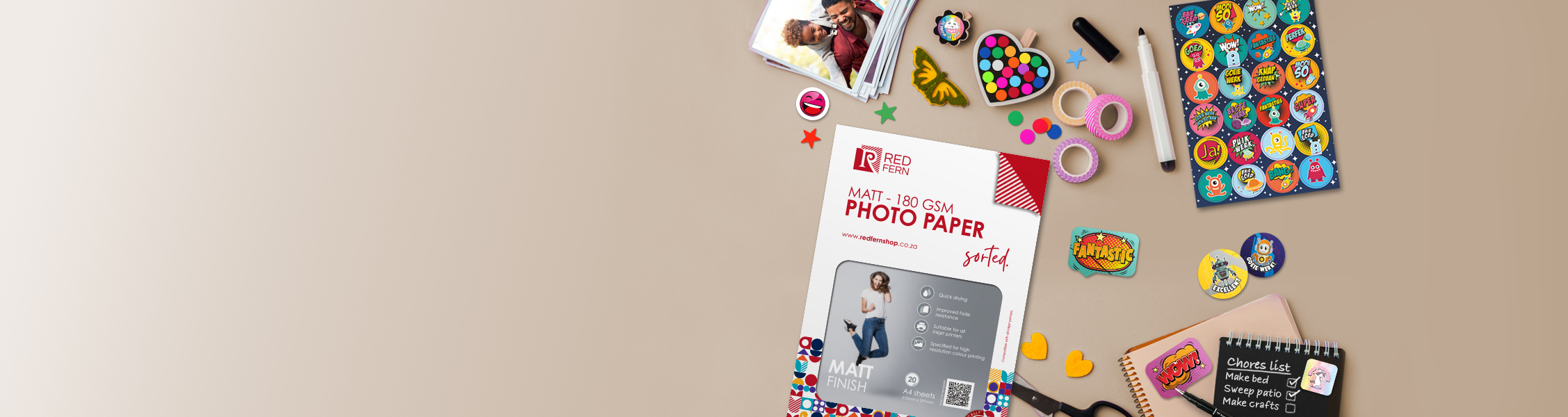 Flat lay of craft supplies on a beige background: photo paper brochure, colorful stickers, tape, scissors, and a notebook cluster, suggesting a scrapbooking scene.